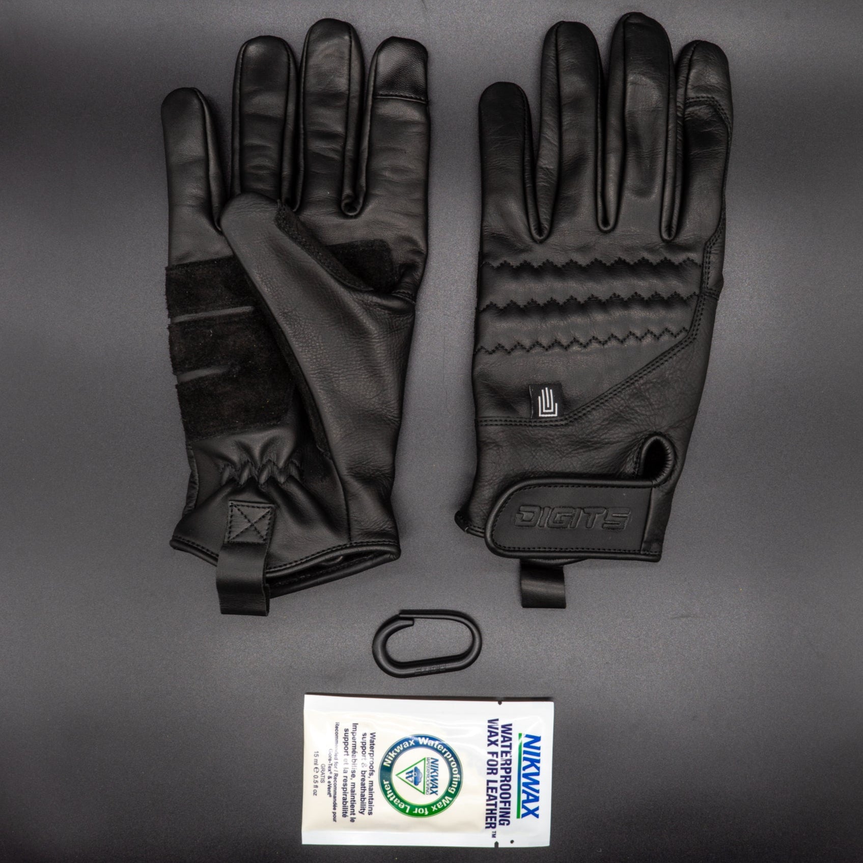 A pair of black, non-insulated leather work gloves with extended cuffs and Velcro wrist closures, displayed palm-up with a company logo on the Velcro strap, alongside a package of Nikwax leather waterproofing.